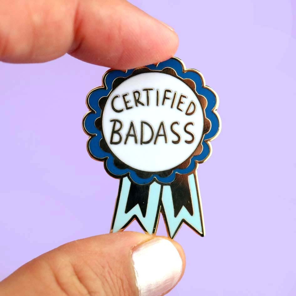 A hard enamel lapel pin being held in a hand. The pin is in the shape of a blue-ribbon award and reads Certified Badass.