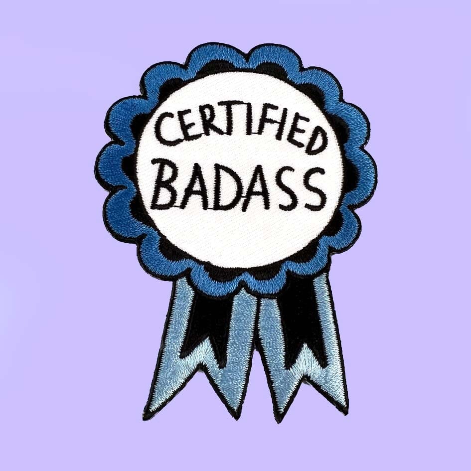 An iron on embroidered patch on Jubly-Umph cardstock against a purple background. The patch is in the shape of a blue-ribbon award and reads Certified Badass. 