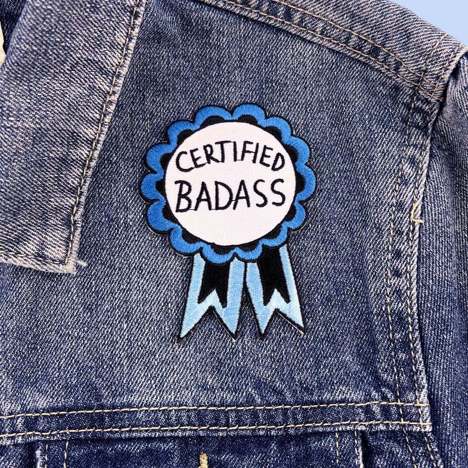 An iron on embroidered patch on a denim jacket. The patch is in the shape of a blue-ribbon award and reads Certified Badass.