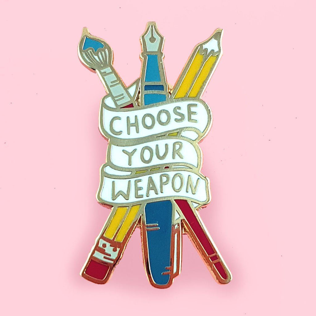 Enamel lapel pin depicting a red paintbrush, a blue fountain pen, and a yellow pencil, all crossed over each other and wrapped in a banner that displays the words 'Choose Your Weapon'. 