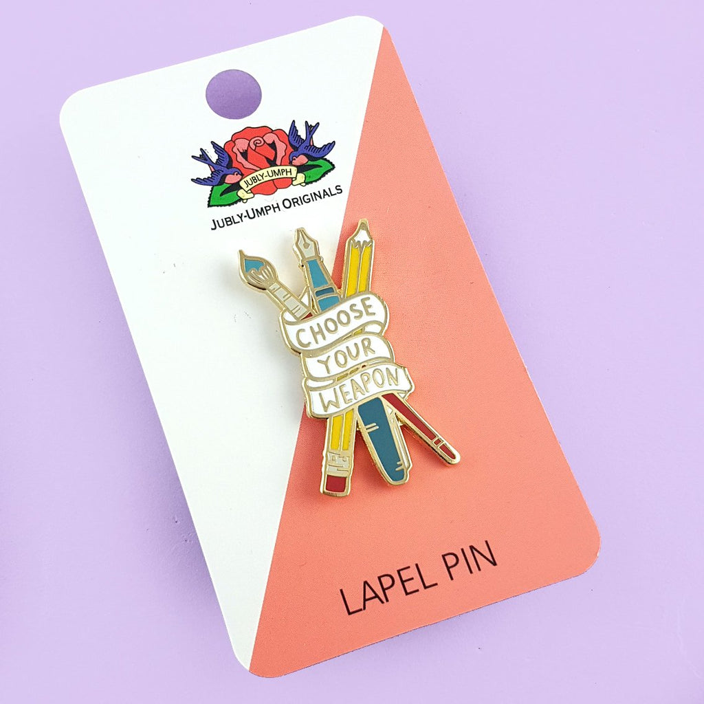 Enamel lapel pin depicting a red paintbrush, a blue fountain pen, and a yellow pencil, all crossed over each other and wrapped in a banner that displays the words 'Choose Your Weapon'. It's pinned to a business card-sized pink and white Jubly-Umph lapel pin card.