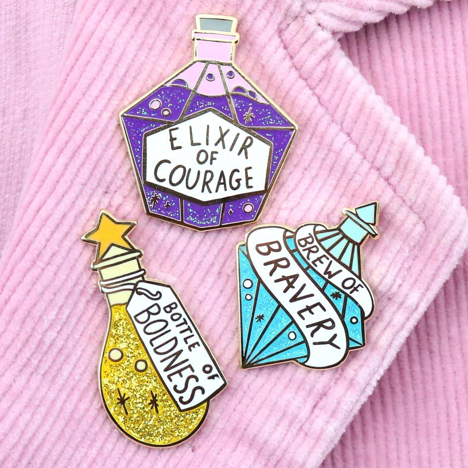 Three hard enamel pins are displayed on a pink jacket. The first pin says Bottle of Boldness, The Second pin says Elixir of Courage and the Third pin says Brew of Bravery.