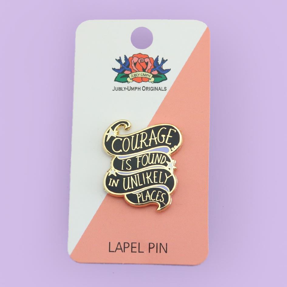 A hard enamel lapel pin on Jubly-Umph cardstock. The pin is black and purple and reads Courage Is Found In Unlikely Places.
