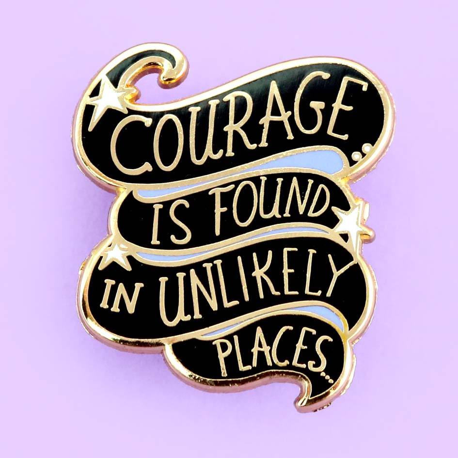 A hard enamel lapel pin on a light purple background. The pin is black and purple and reads Courage Is Found In Unlikely Places.