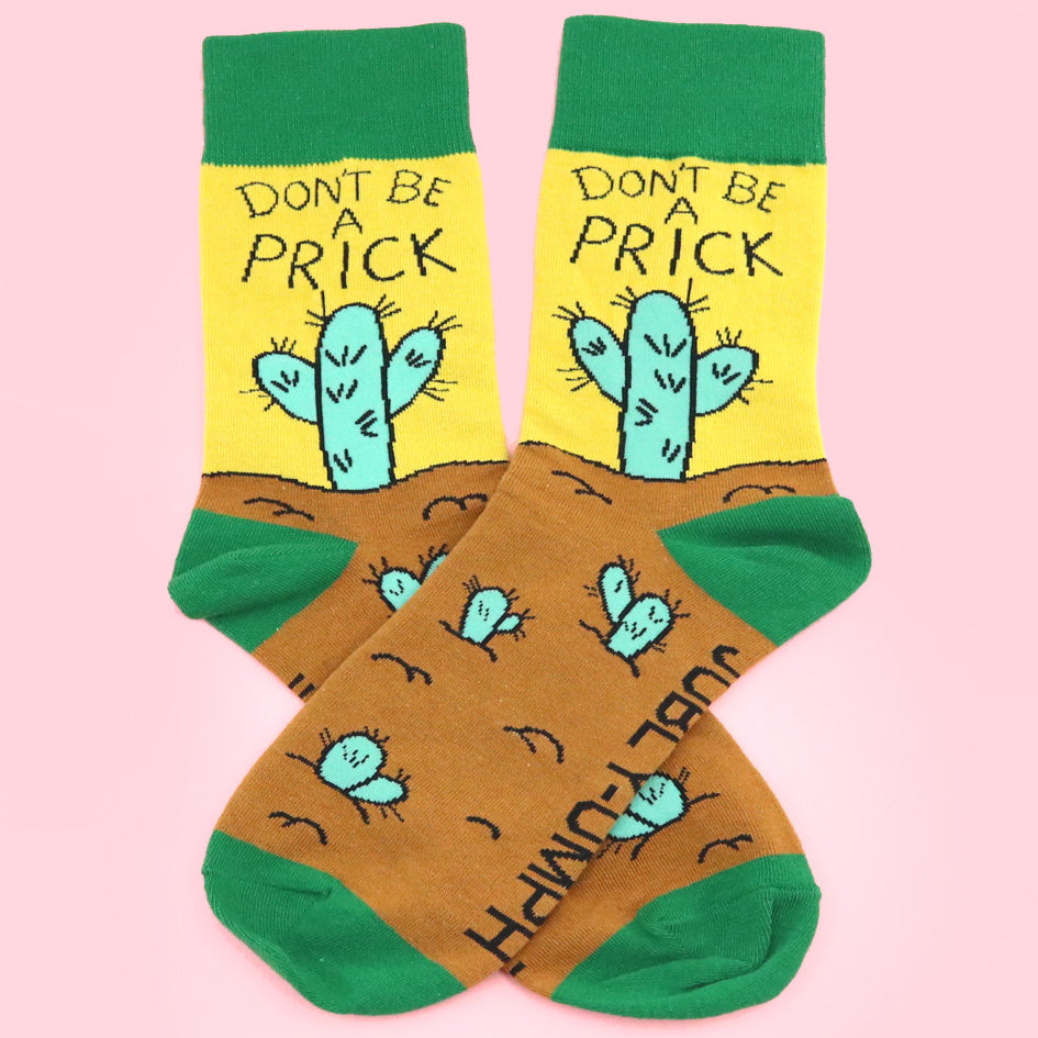 A pair of socks against a pink background. The socks are yellow, green and brown with a teal cactus. The socks read Don't Be A Prick.