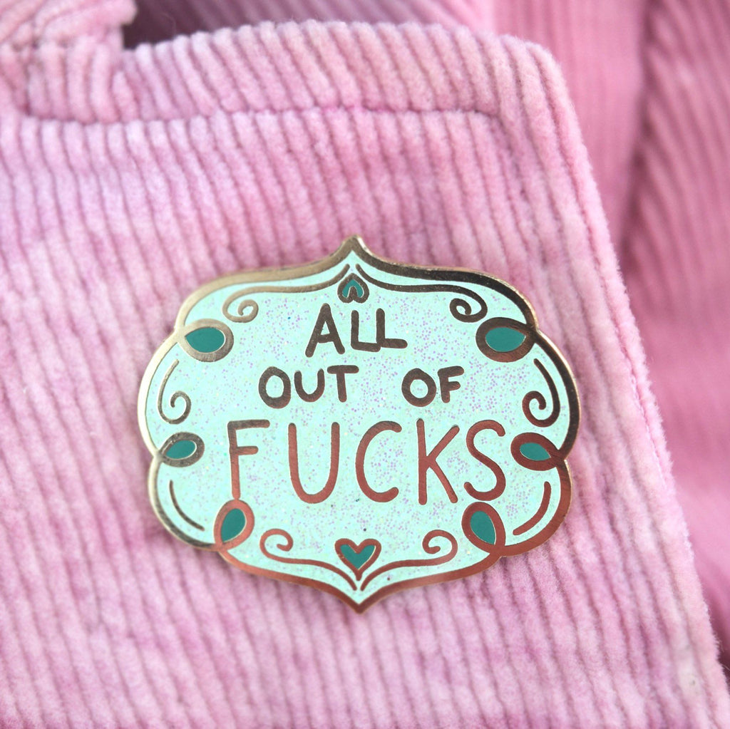 A hard enamel lapel pin being worn on a pink jacket. The pin has blue sparkly enamel and reads All Out Of Fucks.