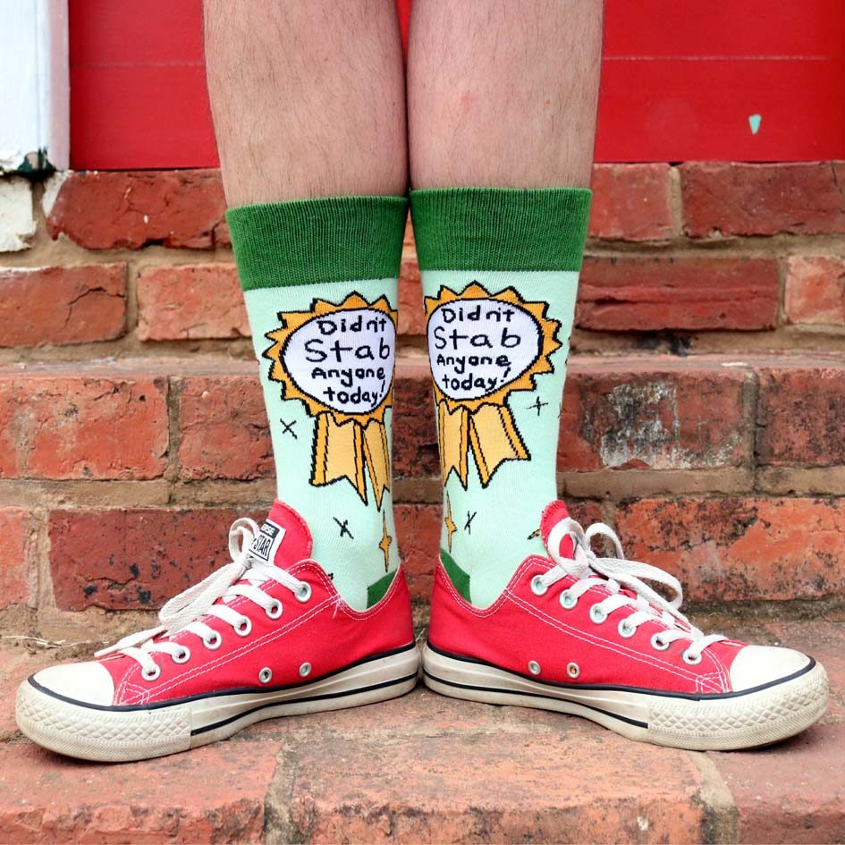 A pair of socks being worn with red shoes standing against a brick background. The socks are green and yellow and read Didn’t Stab Anyone Today!