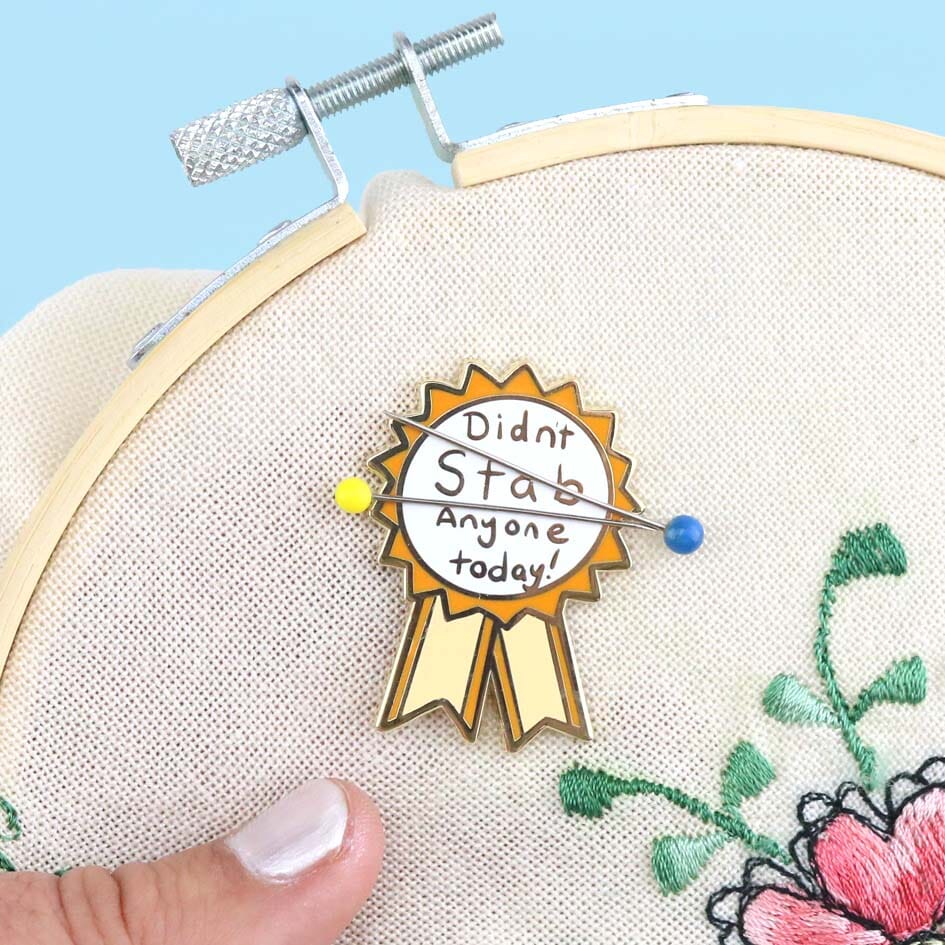 A hard enamel magnetic needle minder displayed on an embroidery hoop with two sewing needles. The pin is in the shape of an award ribbon. The ribbon is yellow and white, and reads Didn’t Stab Anyone Today!