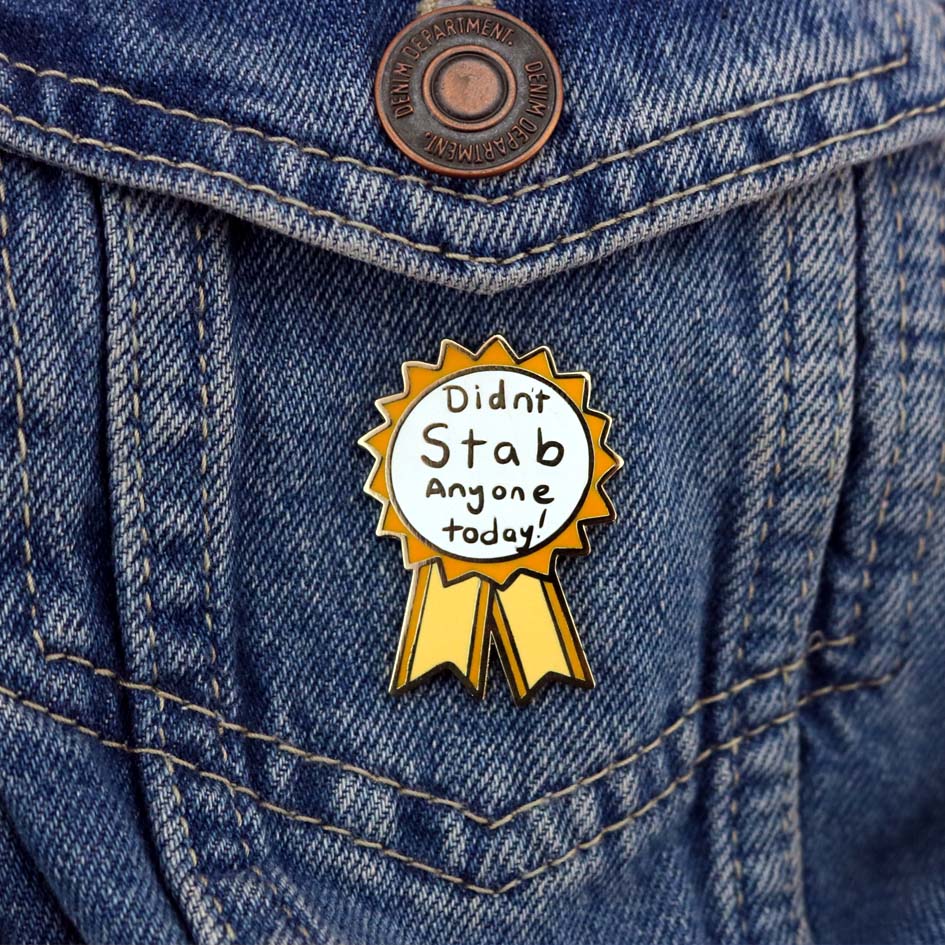 A hard enamel lapel pin on a denim jacket. The pin is in the shape of an award ribbon. The ribbon is yellow and white, and reads Didn’t Stab Anyone Today!