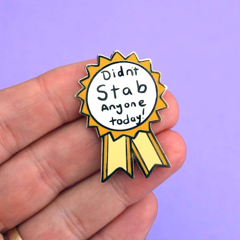 A hard enamel lapel pin being held in a hand. The pin is in the shape of an award ribbon. The ribbon is yellow and white, and reads Didn’t Stab Anyone Today!