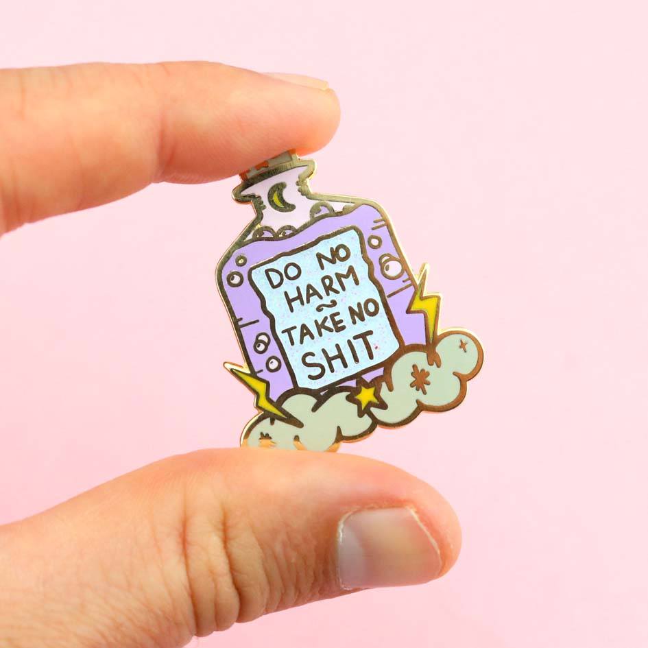 A hard enamel lapel pin being held in hand on a pink background. The pin is purple and in the shape of a bottle with clouds and lightning bolts. The pin reads Do No Harm Take No Shit.