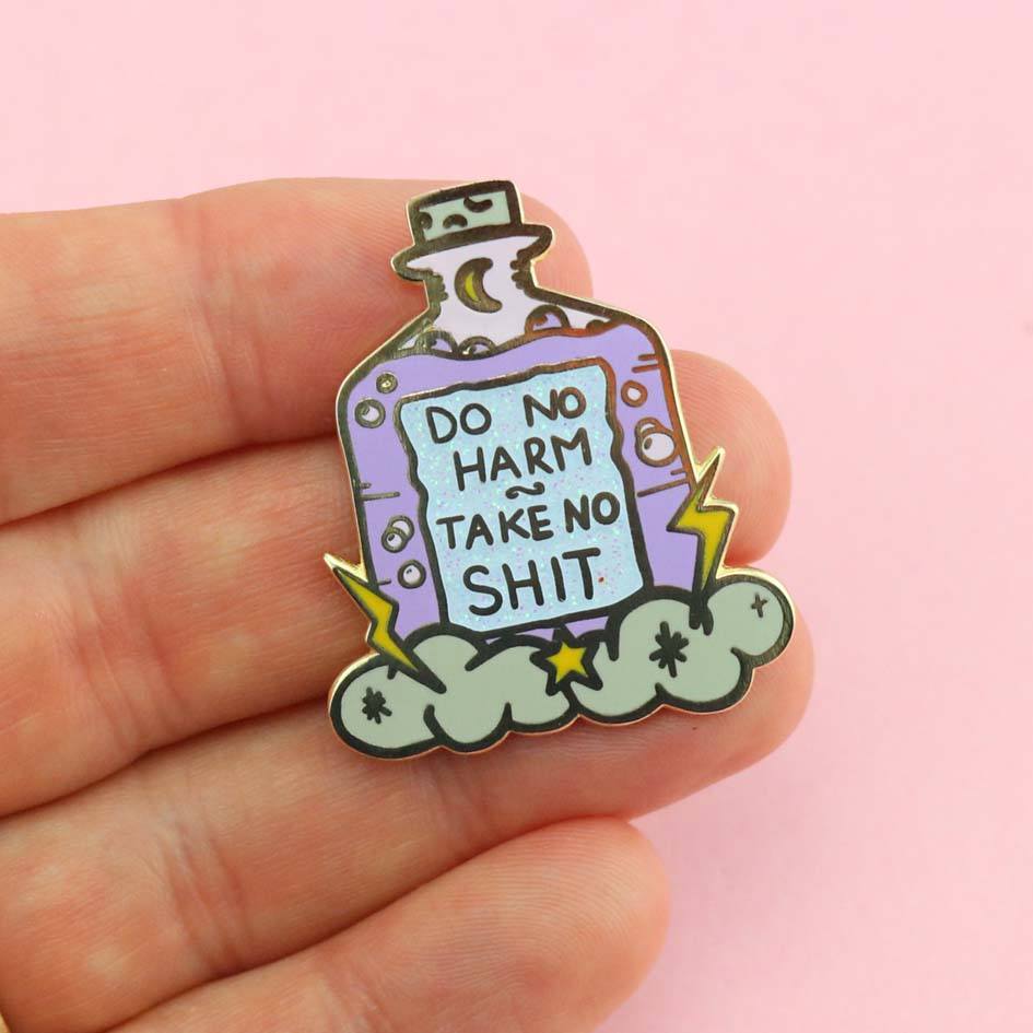 A hard enamel lapel pin being held in hand on a pink background. The pin is purple and in the shape of a bottle with clouds and lightning bolts. The pin reads Do No Harm Take No Shit.