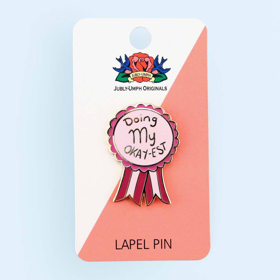 A hard enamel lapel pin on Jubly-Umph cardstock. The pin is in the shape of an award ribbon. The ribbon is dark pink and light pink and reads Doing my Okay-est.