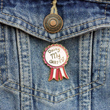 A hard enamel lapel pin on a denim jacket. The pin is in the shape of an award ribbon. The ribbon is dark pink and light pink and reads Doing my Okay-est.