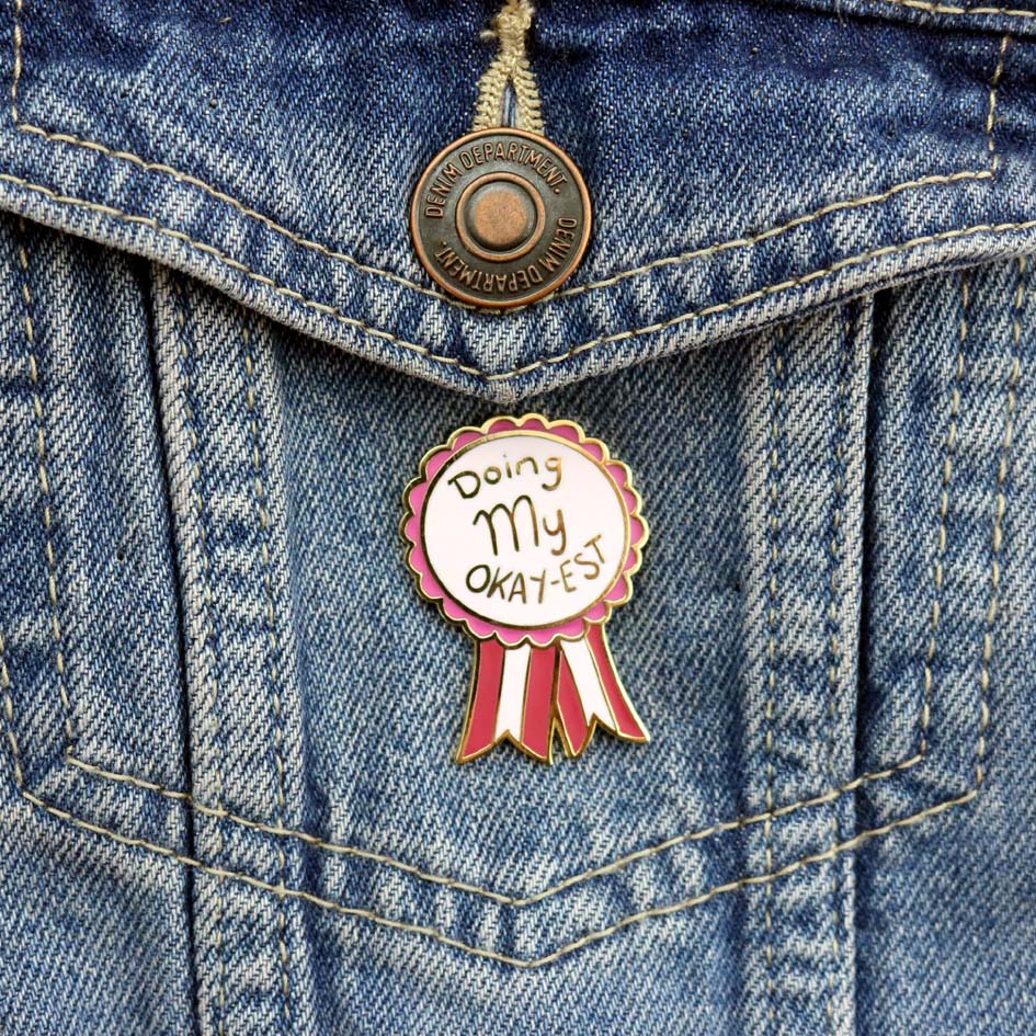 A hard enamel lapel pin on a denim jacket. The pin is in the shape of an award ribbon. The ribbon is dark pink and light pink and reads Doing my Okay-est.