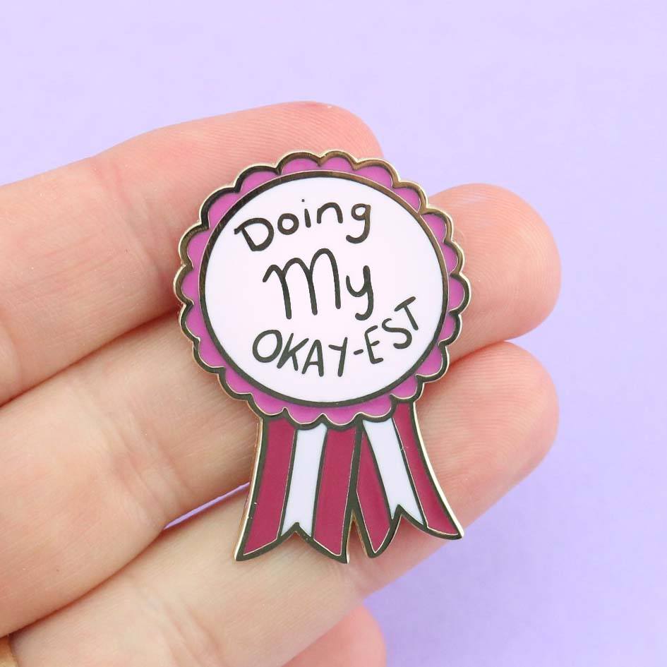 A hard enamel lapel pin being held in a hand. The pin is in the shape of an award ribbon. The ribbon is dark pink and light pink and reads Doing my Okay-est.