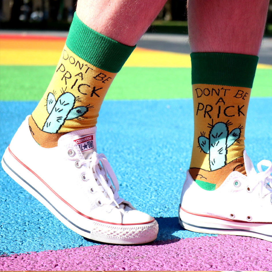 A pair of socks being worn with white shoes with a rainbow background. The socks are yellow, green and brown with a teal cactus. The socks read Don't Be A Prick.