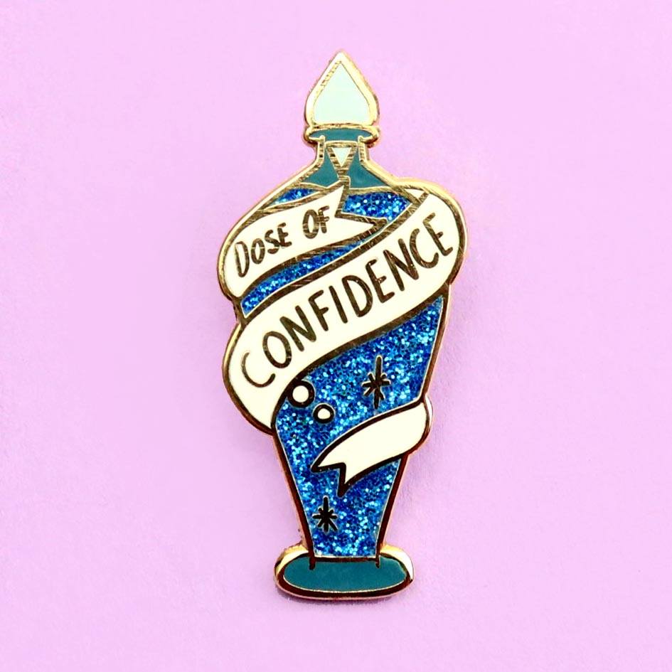 A hard enamel lapel pin on a purple background. The pin is in the profile of a bottle with dark blue glitter. There is a white ribbon around the bottle that reads Dose Of Confidence.
