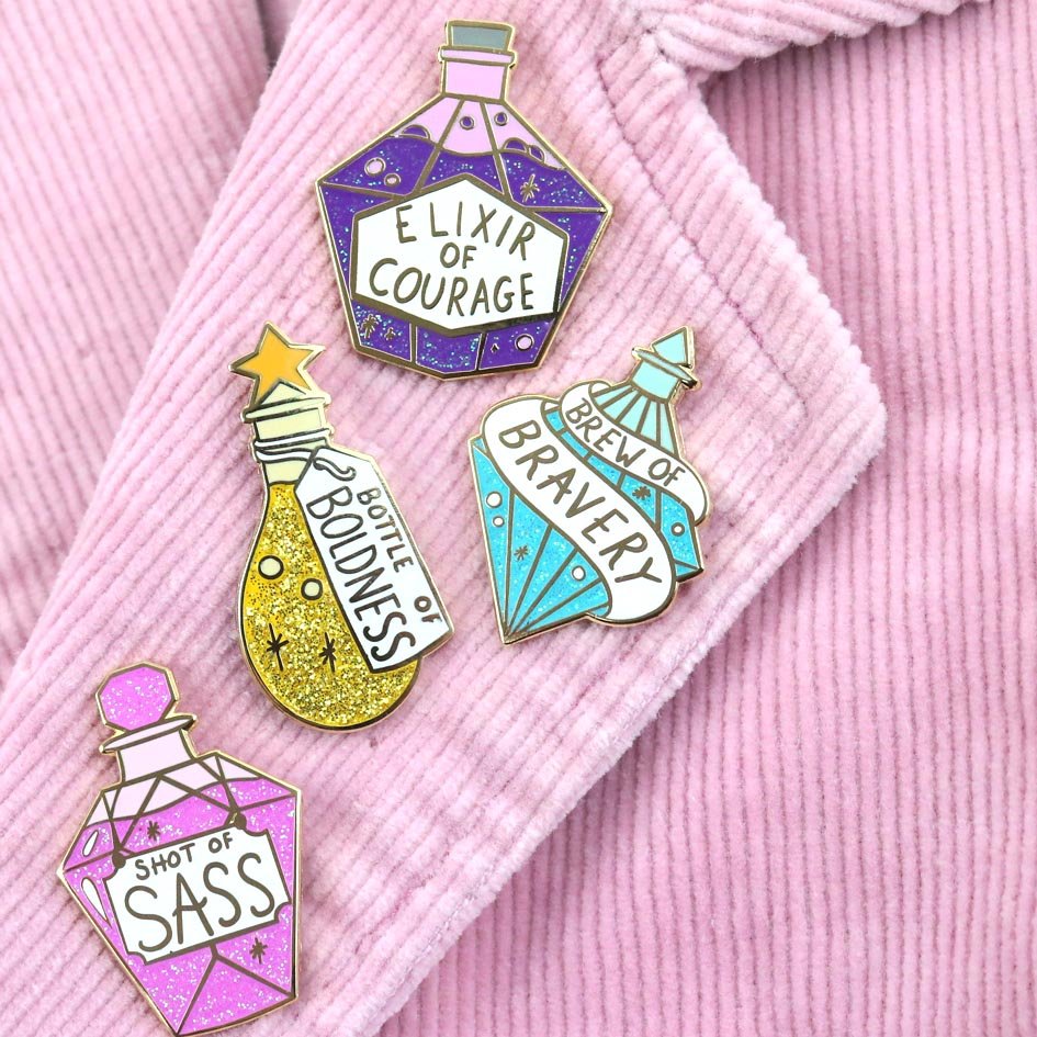Four hard enamel pins are displayed on a pink jacket. The first pin says Bottle of Boldness, The Second pin says Elixir of Courage, the Third pin says Brew of Bravery and fourth pin says Shot of Sass.