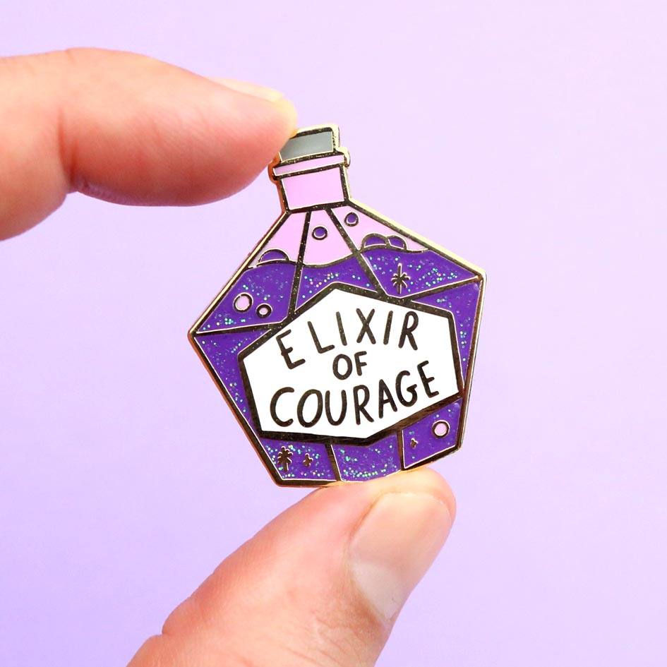A hard enamel pin being held in hand on a purple background. The pin is in the shape of a bottle with purple glitter. The pin reads Elixir Of Courage.