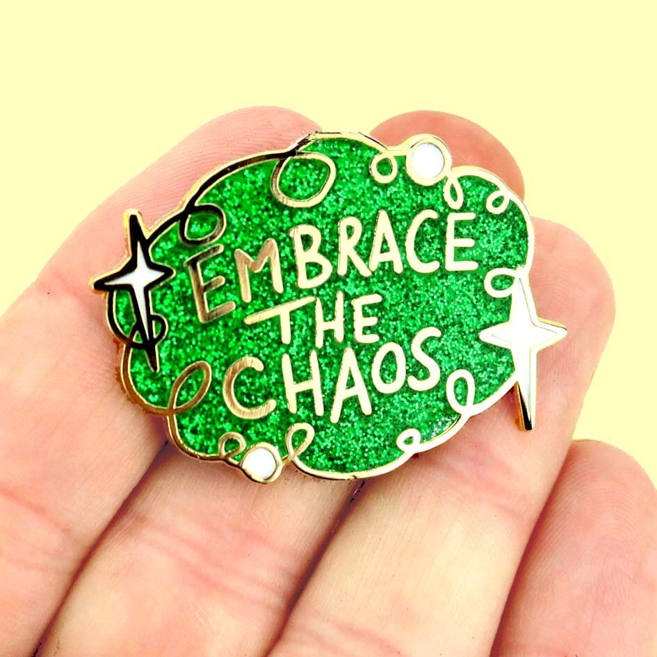 A hard enamel lapel pin being held in the hand on a yellow backround. The pin is green glitter with white stars and reads Embrace the Chaos.