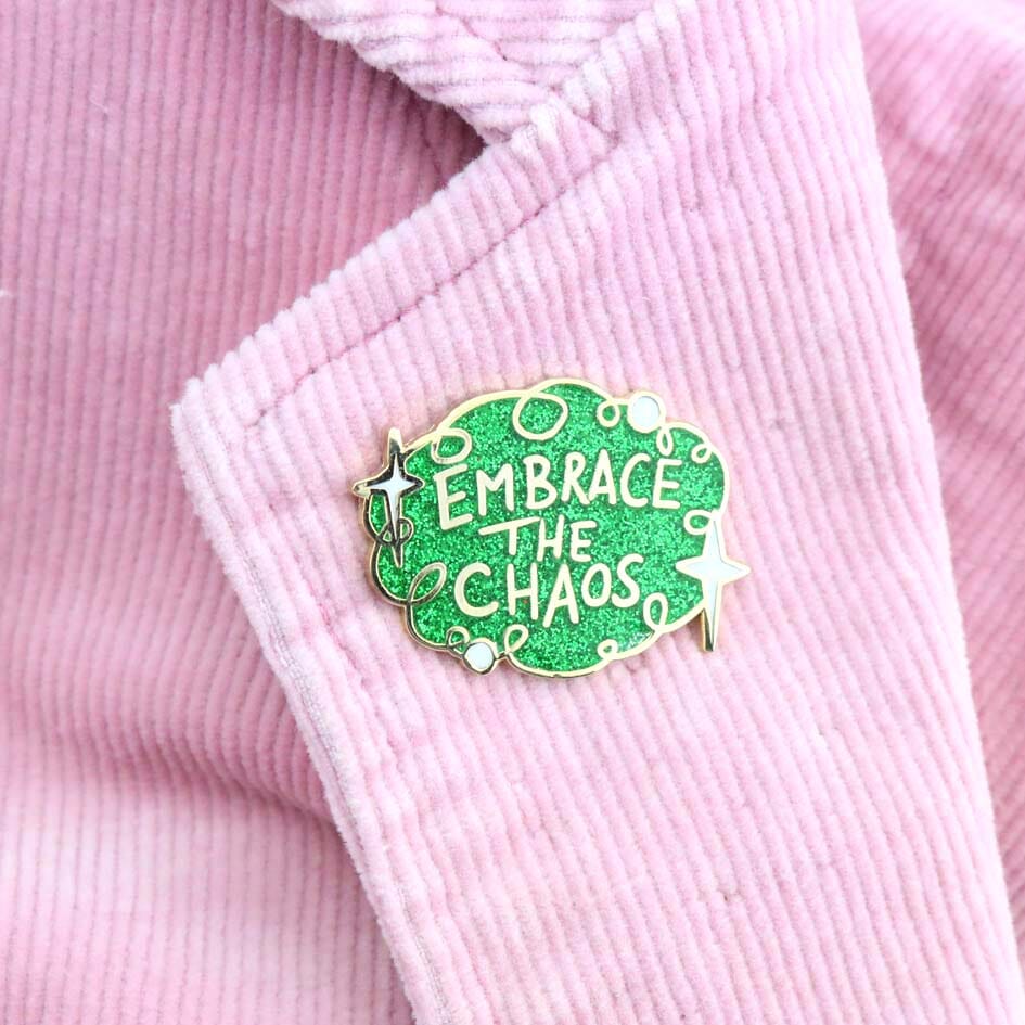 A hard enamel lapel pin on a pink jacket. The pin is green glitter with white stars and reads Embrace the Chaos.