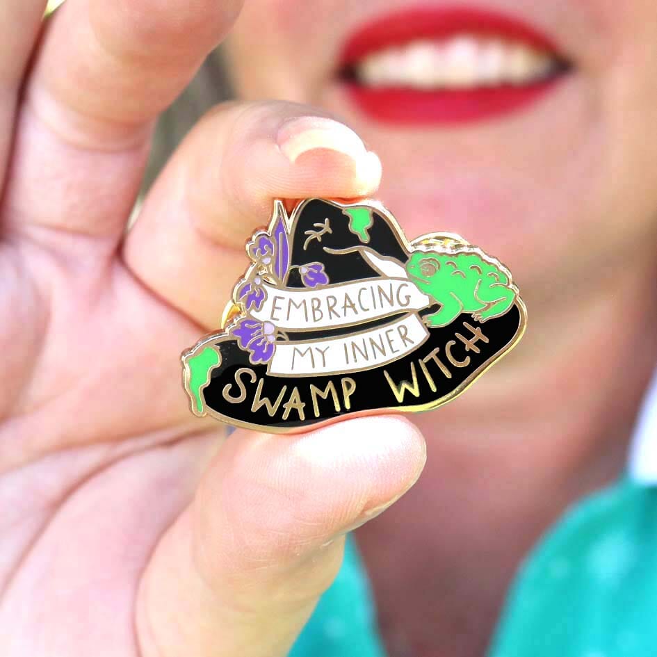 A hard enamel pin being held in hand. The pin is black in the shape of a witches hat. The hat reads Embracing My Inner Swamp Witch.