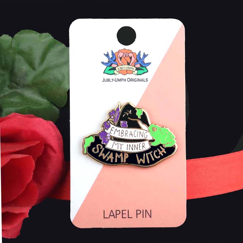A hard enamel pin on Jubly-Umph card stock. The pin is black in the shape of a witches hat. The hat reads Embracing My Inner Swamp Witch.