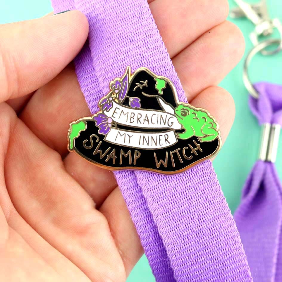 A hard enamel lapel pin being held in the hand and attached to a purple lanyard. The pin is black in the shape of a witches hat. The hat reads Embracing My Inner Swamp Witch.