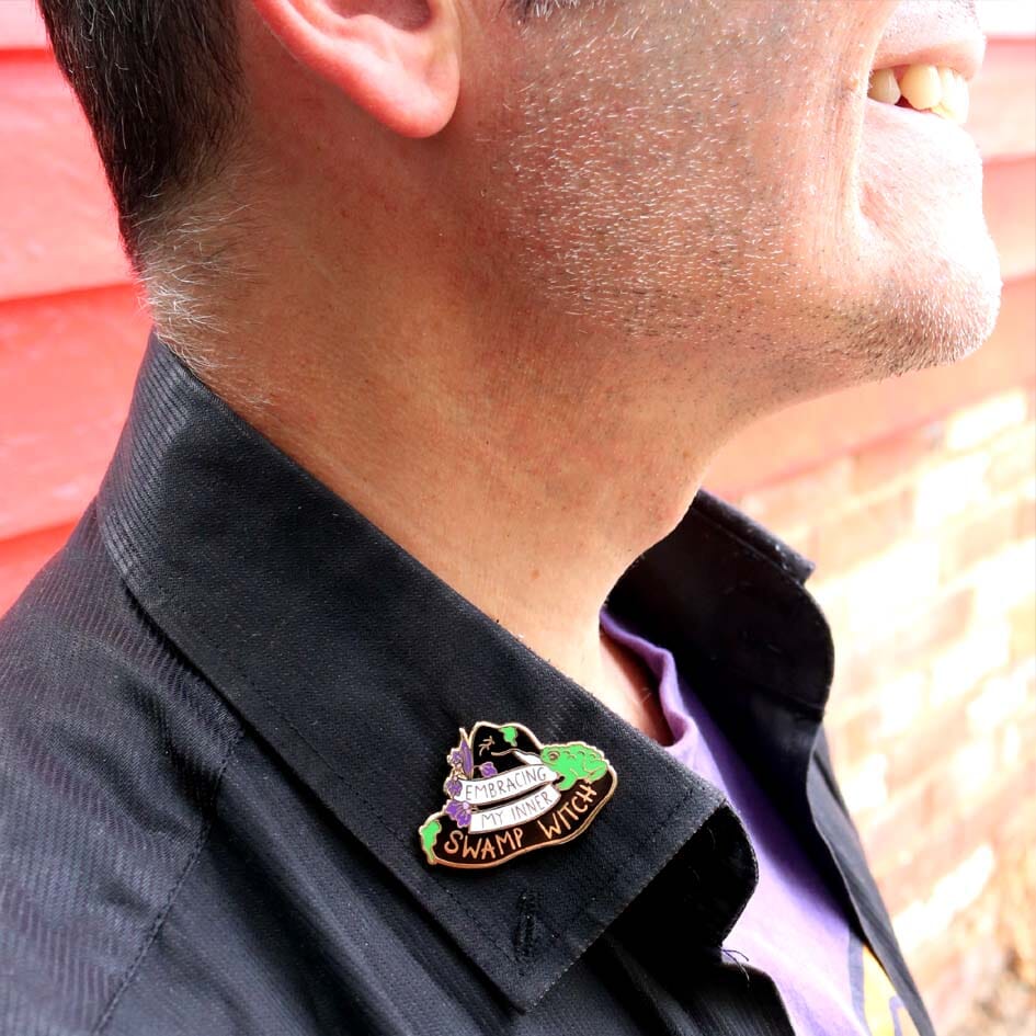 A hard enamel pin being worn on a black shirt. The pin is black in the shape of a witches hat. The hat reads Embracing My Inner Swamp Witch.