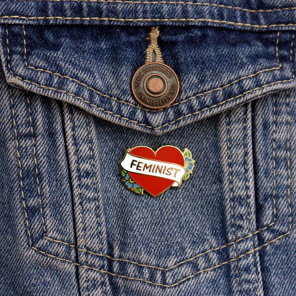 A hard enamel pin on a denim jacket. The pin is in the shape of a red heart and reads Feminist.