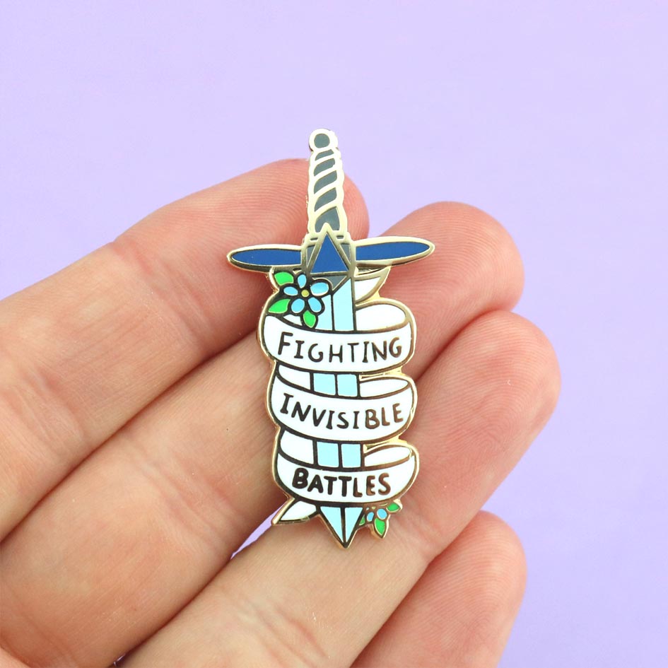 A hard enamel pin held in the hand. The pin is in the shape of a dagger and reads Fighting Invisible Battles.