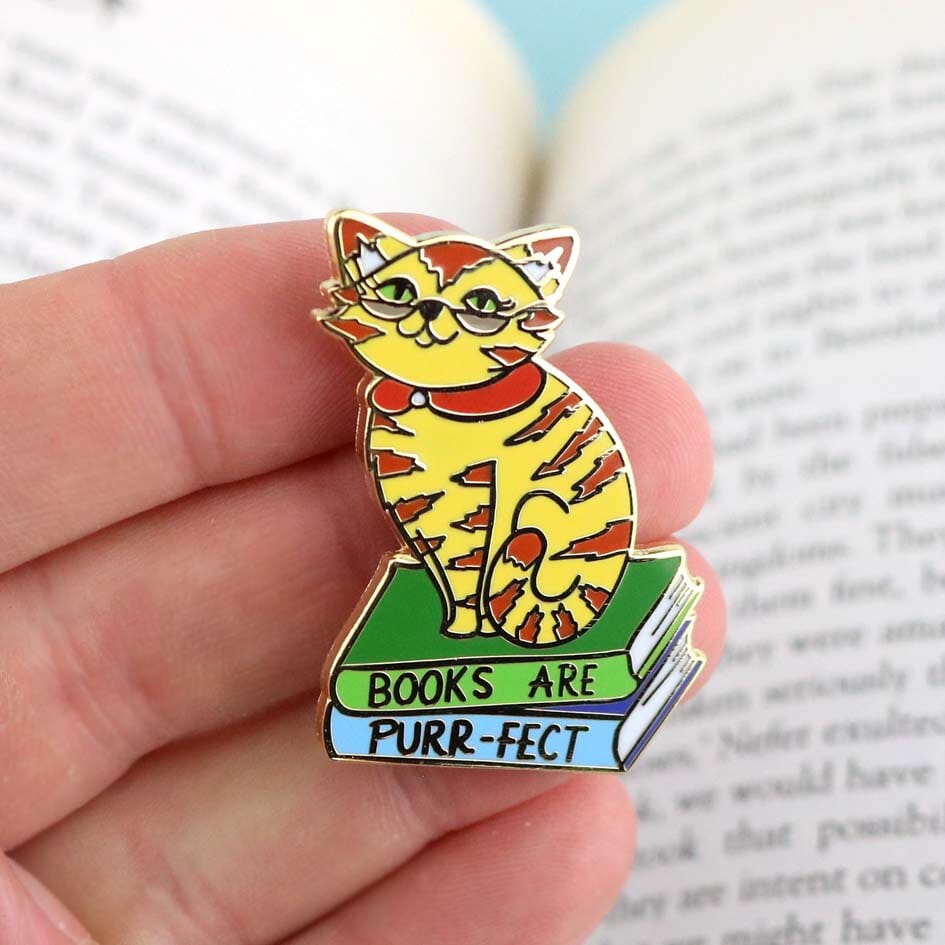 A hard enamel lapel pin being held in a hand. The pin says Books Are Purr-fect with a cat sitting on books.