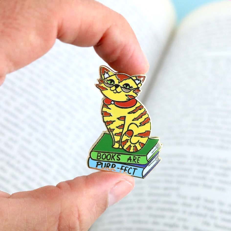 A hard enamel lapel pin being held in a hand. The pin says Books Are Purr-fect with a cat sitting on books.