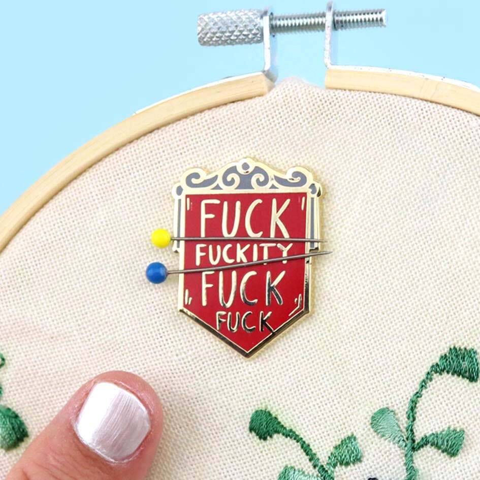 A hard enamel magnetic needle minder on a embroidery hoop with two needles. The pin is in the shape of a red shield and reads Fuck Fuckity Fuck Fuck.