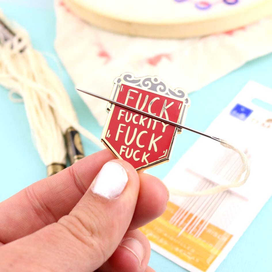 A hard enamel magnetic needle minder held in the hand with a needle. The pin is in the shape of a red shield and reads Fuck Fuckity Fuck Fuck.