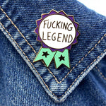 A hard enamel lapel pin being displayed on a denim jacket. The pin is in the shape of a teal and purple ribbon award and reads Fucking Legend.