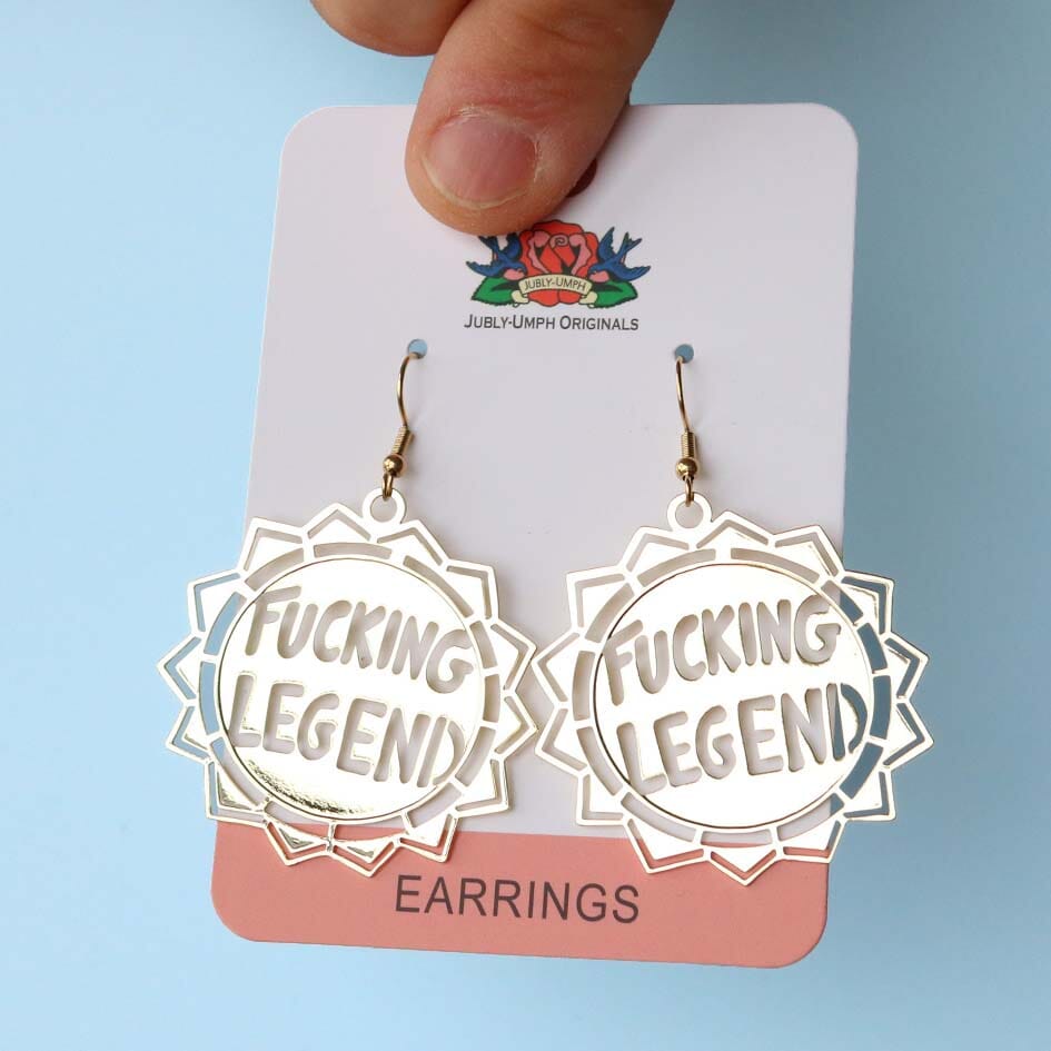 A pair of dangle brass earrings displayed on Jubly-Umph card stock. The earrings read Fucking Legend.