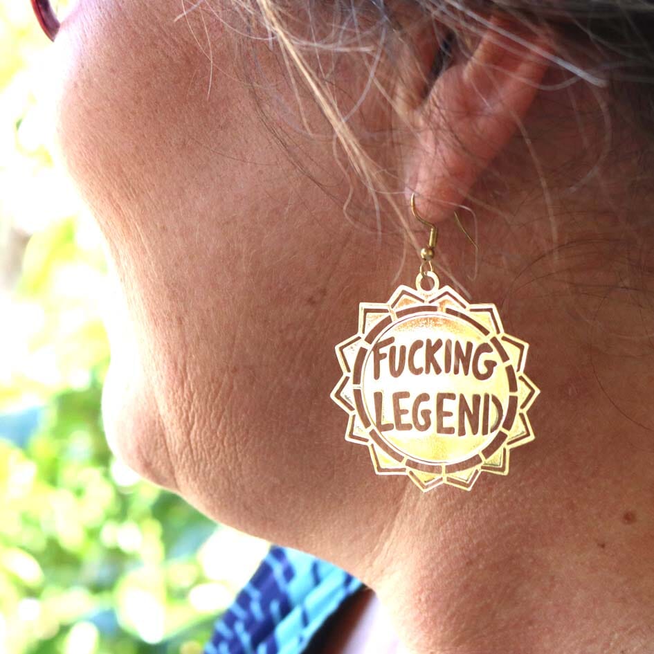 A single brass earring being worn by a model. The earring reads Fucking Legend.