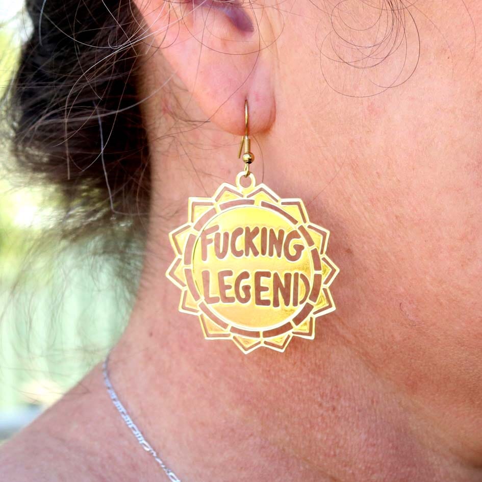 A single brass earring being worn by a model. The earring reads Fucking Legend.
