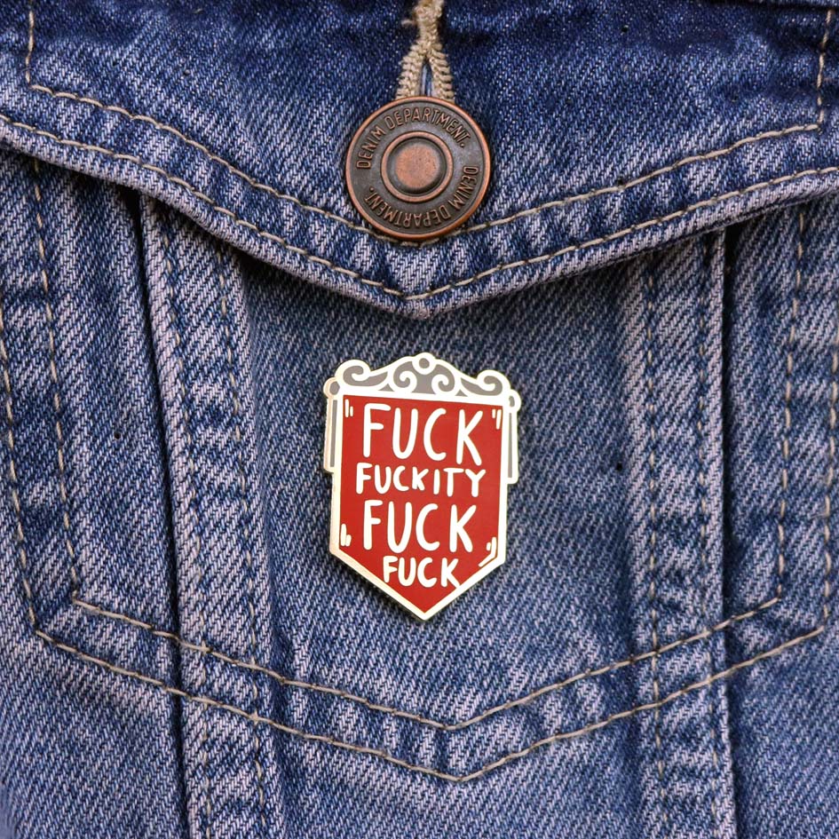A hard enamel pin on denim jacket. The pin is in the shape of a red shield and reads Fuck Fuckity Fuck Fuck.