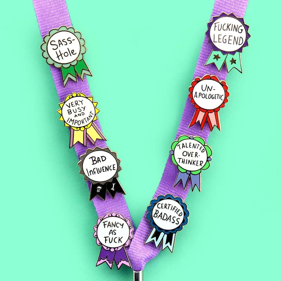 Eight hard enamel pins are attached to a lanyard. They are each in the shape of an award ribbon. They read Sass-Hole, Very Busy and Important, Bad Influence, Fancy As Fuck, Fucking Legend, Un-apologetic, Talented Overthinker, Certified Badass.