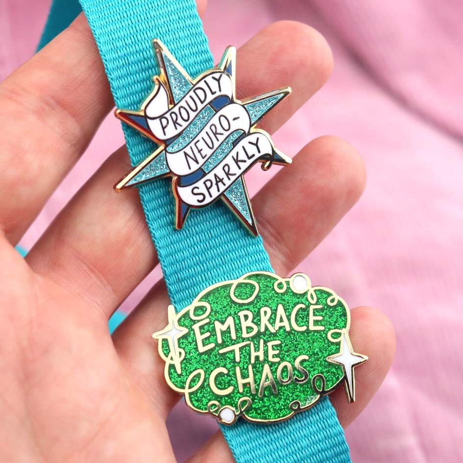 Two hard enamel lapel pins being held in the hand and attached to a blue lanyard. The first pin is in the shape of a blue star and reads Proudly Neuro-Sparkly. The second pin is green glitter with white stars and reads Embrace the Chaos.