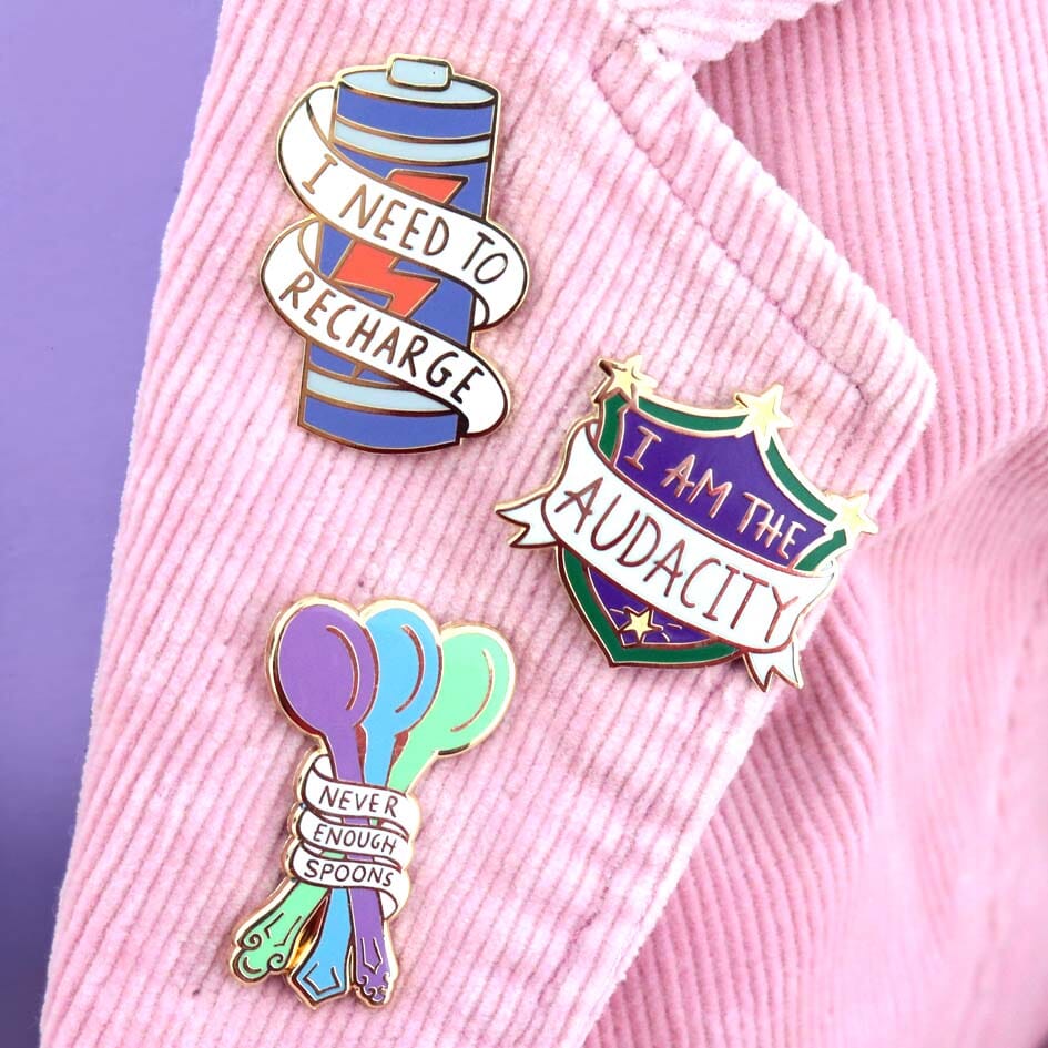 Hard enamel lapel pins on a pink jacket. The lapel pins read I Need To Recharge, I Am The Audacity and Never Enough Spoons.