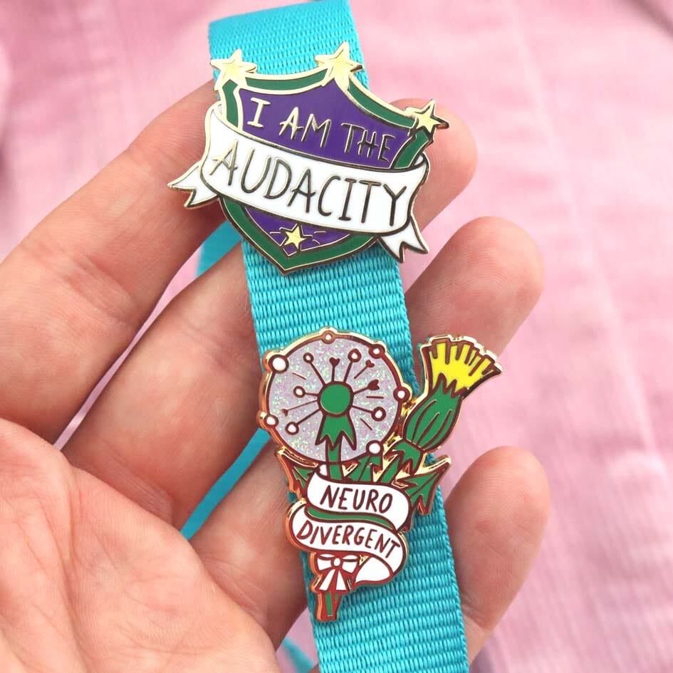 A hard enamel lapel pin being held in a hand on a blue lanyard with another Jubly-Umph pin. The pin is in the shape of a shield. The lapel pin reads I am the Audacity.