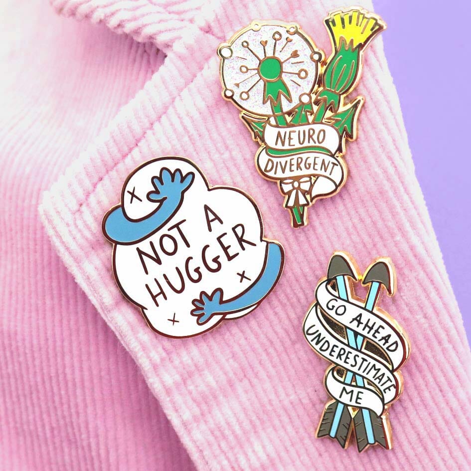 A hard enamel lapel pin being displayed alongside other Jubly-Umph pins. The pin is in the shape of in the shape of two arrows and reads Go Ahead Underestimate Me.