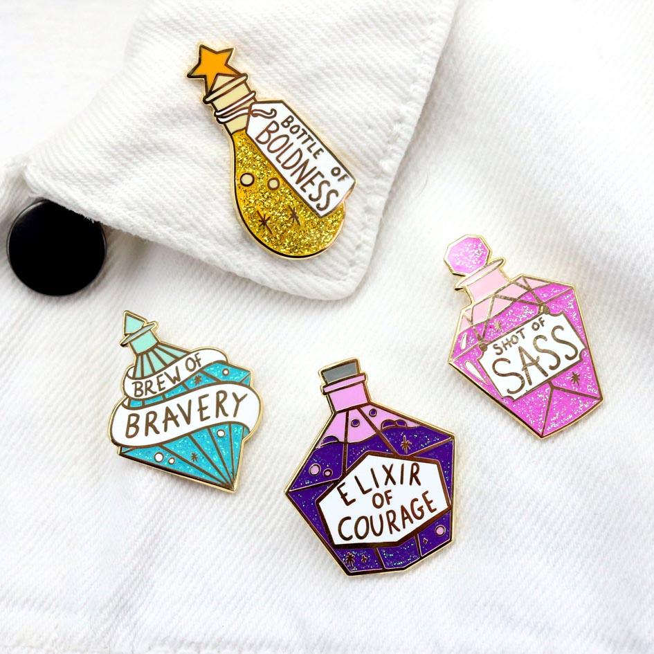 A hard enamel lapel pins are pinned on a white jacket. All the pins are in the shape of an individual stylised bottle. The main bottle is in the shape of a bottle with purple glitter. The pin reads Elixir Of Courage. The other pins read, Shot of Sass, Bottle of Boldness and Brew of Bravery.