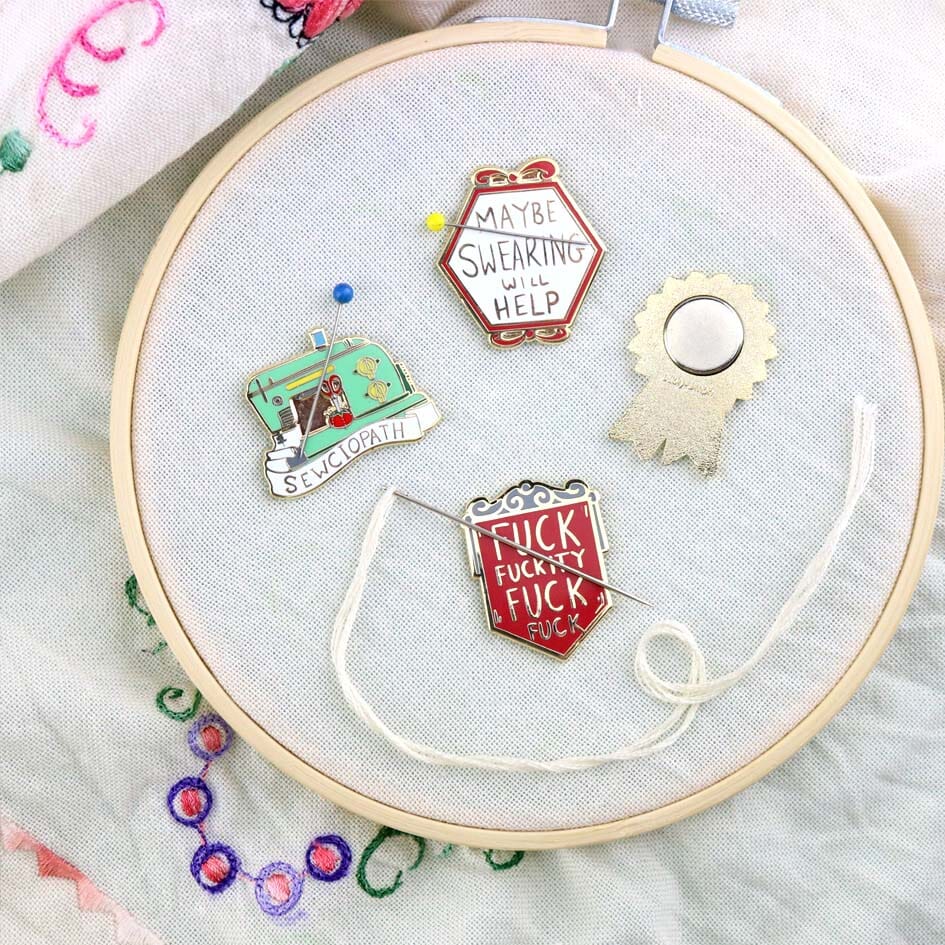 A hard enamel magnetic needle minder on an embroidery hoop with other Jubly-Umph needle minders. The pin is in the shape of a red shield and reads Fuck Fuckity Fuck Fuck.