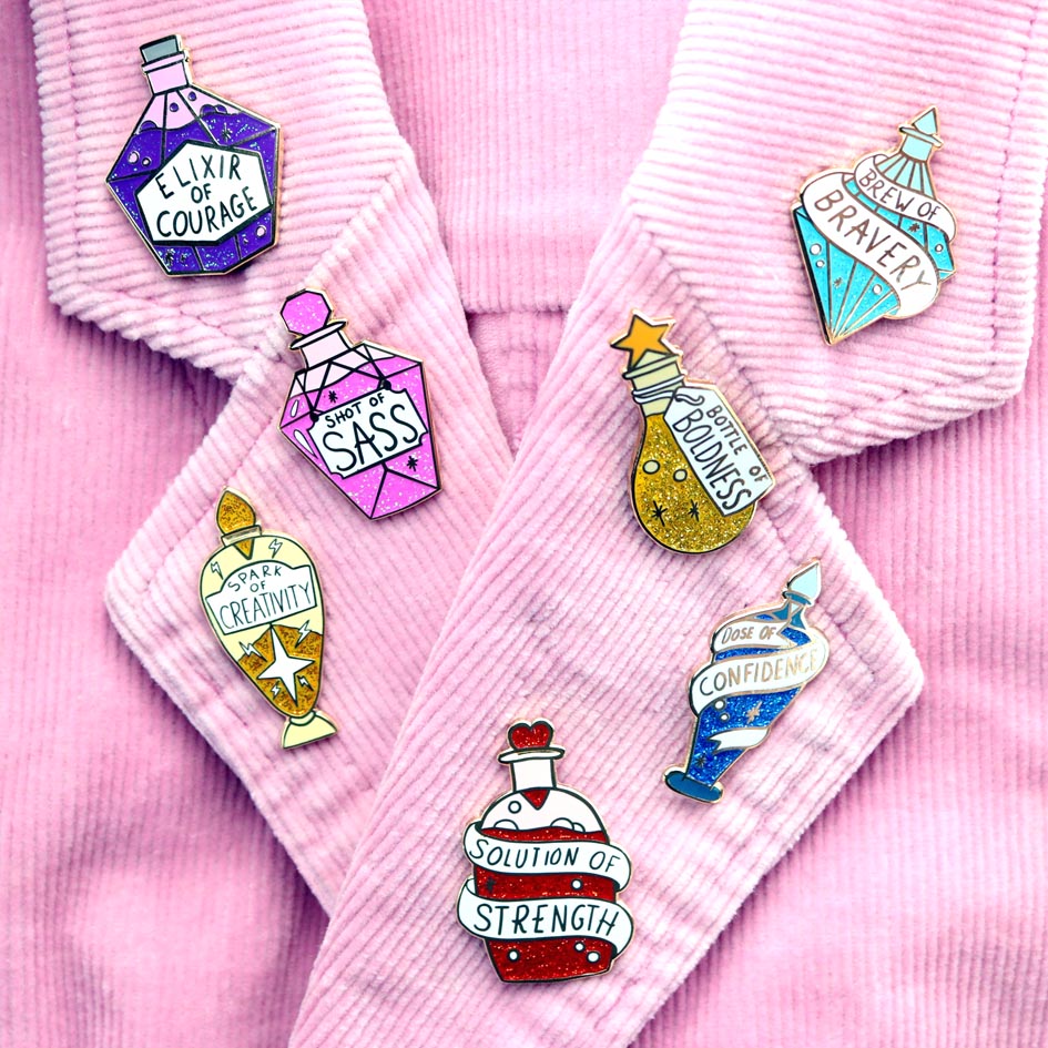 A hard enamel lapel pins are pinned on a pink jacket. All the pins are in the shape of an individual stylised bottle. The pin is in the profile of a bottle with dark blue glitter. There is a white ribbon around the bottle that reads Dose Of Confidence. The other pins read, Elixer of Courage, Shot of Sass, Spark of Creativity, Solution of Strength, Bottle of Boldness and Brew of Bravery.