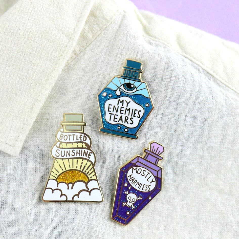 Three hard enamel pins are displayed on a white linen shirt. The first pin says Bottled Sunshine, The Second pin says My Enemies Tears and the Third pin says Mostly Harmless.